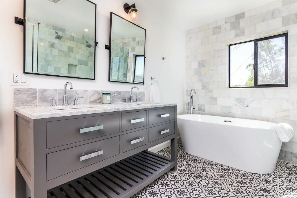 Modern bathroom featuring a gray double vanity with marble countertop, two mirrors, and a freestanding tub, complemented by patterned floor tiles and natural light from a window.