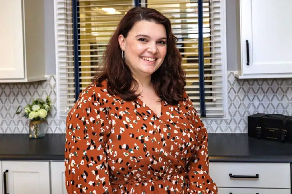 Smiling woman in a patterned dress standing in a modern kitchen with stylish cabinetry and decorative accents, representing design expertise at Dartmouth Building Supply's Design Center.